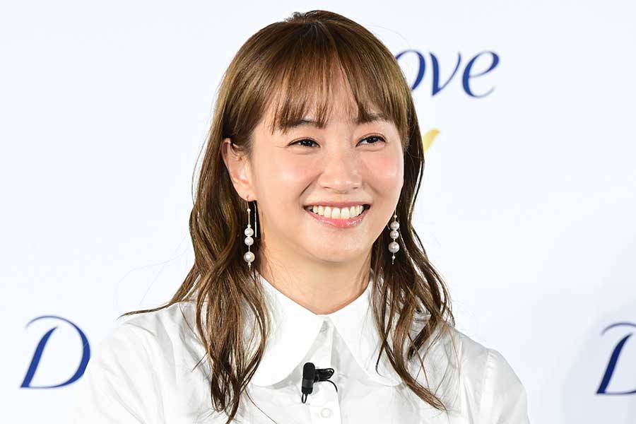 白いシャツ姿で笑顔を向ける藤本美貴が、美容ブランドのロゴ前で撮影されたイベント写真。ピアスとナチュラルメイクが映える、ママタレントとしてのイメージや年収解説記事のアイキャッチに最適な画像。