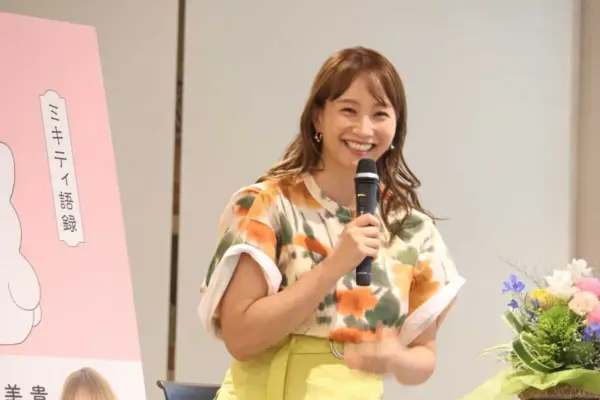 カラフルなトップス姿でマイクを持ち、トークイベントで笑顔を見せる藤本美貴の写真。著書のパネルが横に並び、ママタレントとしての活動や年収記事の挿絵に適したカット。