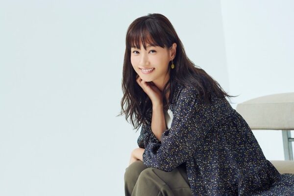 椅子に座り、手を頬に当てて柔らかくほほ笑む藤本美貴のポートレート。落ち着いた衣装と背景で、経歴紹介やプロフィール、年収まとめ記事の上部に使いやすい宣材風写真。