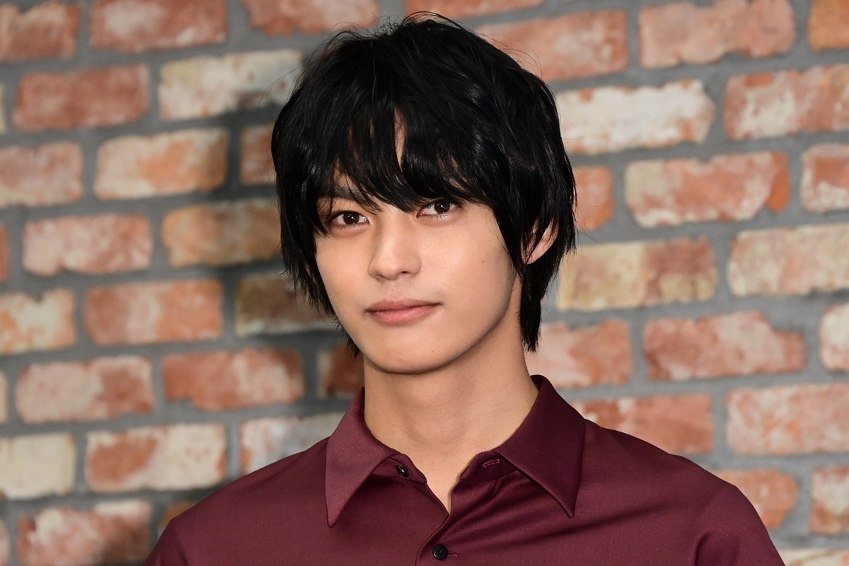 レンガ壁を背景にボルドー色のシャツを着た若い男性俳優が正面を見つめる上半身アップのアイキャッチ用ポートレート写真