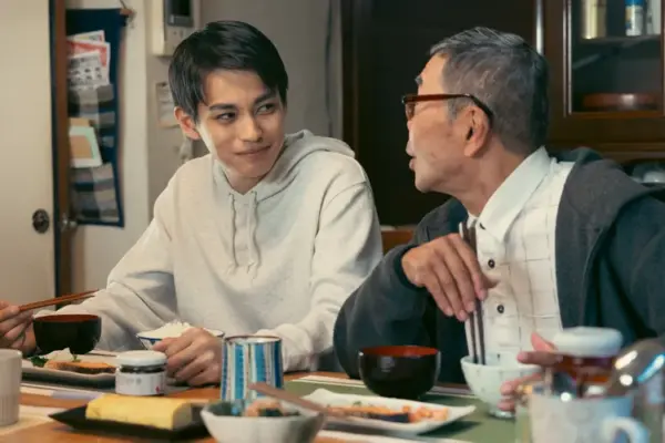 和食の食卓でパーカー姿の若い男性俳優と年配男性が向かい合い笑顔で会話している心温まる家族団らんシーンの写真