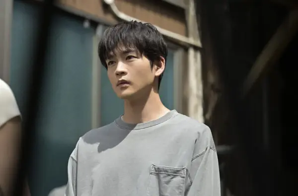 グレーのスウェットを着た若い男性が屋外でまっすぐ前を見つめる横位置写真。等身大の雰囲気が伝わるスチールで、昨年のドラマ出演や俳優の仕事量・収入の変化を説明する記事向けイメージ。