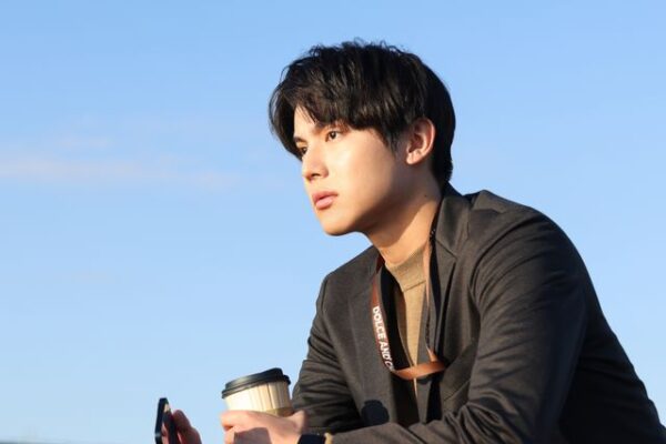 コーヒーを手に青空の下で遠くを見つめる男性俳優の横顔ショット（インタビュー風）