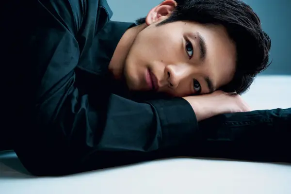黒いシャツ姿の男性が床に横たわり、カメラを見つめるアップのポートレート写真。落ち着いた表情が印象的で、俳優の経歴やプロフィール、インタビュー記事に適したビジュアル。