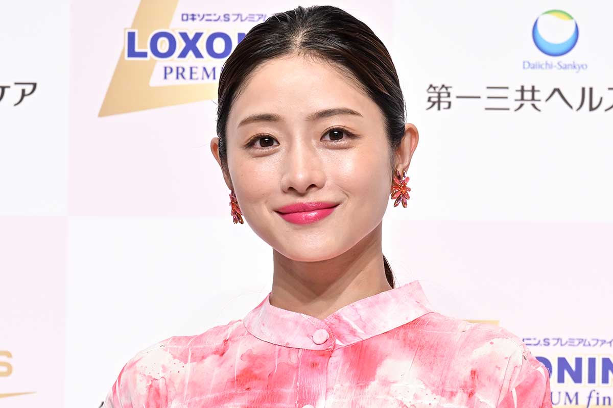 イベント登壇時の石原さとみのクローズアップ。ピンクの衣装とイヤリング、背後に医薬品ブランドのボードが見える宣伝会見風景。