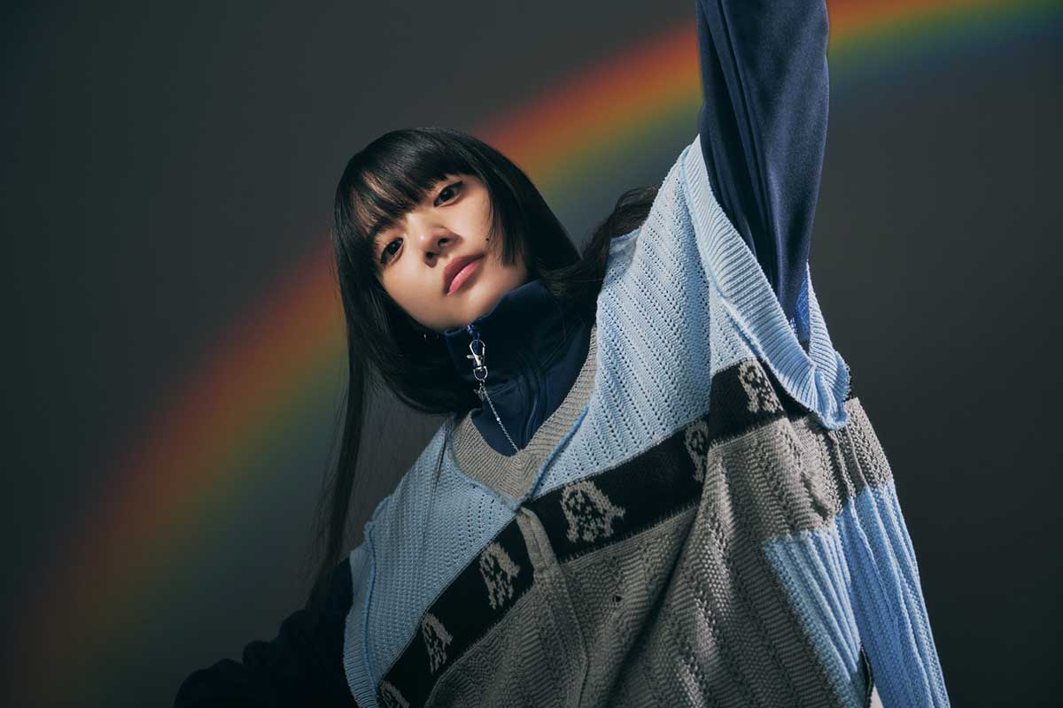 虹を背景にニットベスト姿でポーズを取るあいみょんのビジュアル写真（個性的なファッションとブランディングが年収アップに繋がる様子をイメージ）