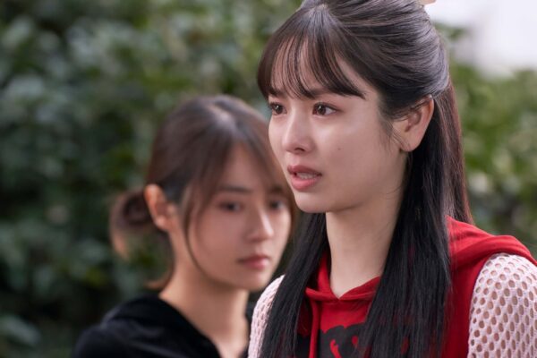 横田真悠が赤いパーカー姿で涙をこらえながら真剣な表情を見せ、後ろには共演者がぼかして写っているドラマの感情的な場面カット