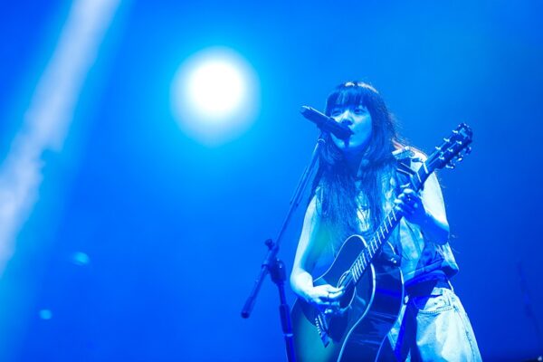 青い照明の中でギターを弾きながら熱唱するあいみょんのライブ写真（音楽活動と年収の関係を伝えるステージショット）
