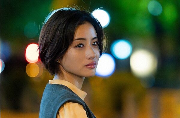 夜景のボケを背景に、ショートヘアの石原さとみが横顔で振り向くポートレート。柔らかな照明が表情を際立たせる。