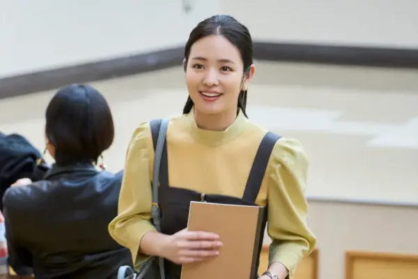 横田真悠が黄緑色のブラウスとジャンパースカート姿でノートを抱え、大学キャンパス風の場所で笑顔を見せているドラマシーンの写真