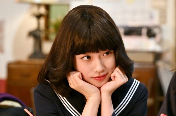 セーラー服姿の若い女性が両手で頬を支え、室内で横目に微笑むシーン写真