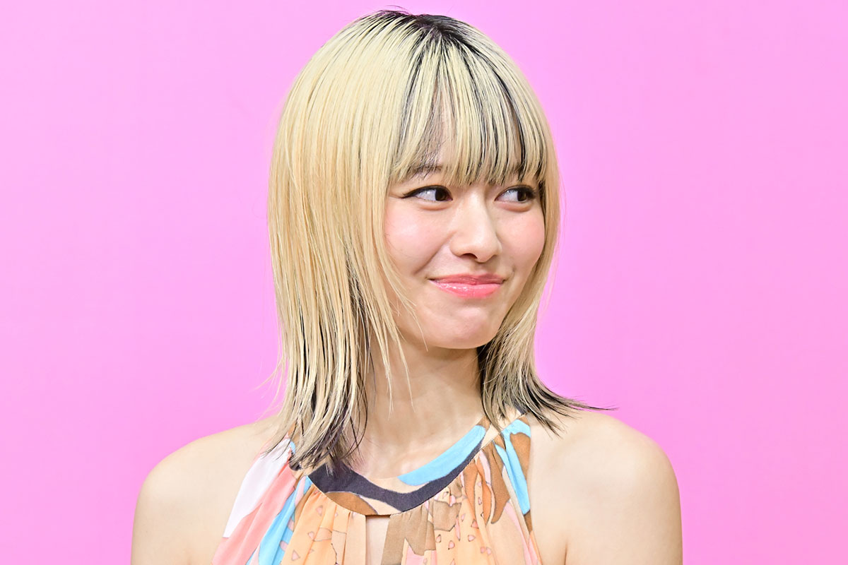 ピンク色の背景をバックに笑顔を浮かべる金髪ボブヘアの女性タレントの上半身アップ・年収解説コラムのアイキャッチに使える明るいイメージ写真