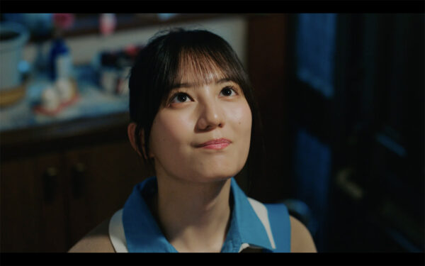 制服風の衣装を着た若い女性アイドルが室内で見上げてほほえむドラマシーン風カット