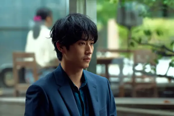屋外のカフェ風の背景で、青いスーツ姿の若い男性が横を向いて座るドラマのワンシーン画像