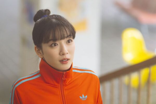 オレンジ色のジャージを着た若い女性が驚いた表情で見上げる映画スチール。スポーティーな装いと明るい色味が印象的。
