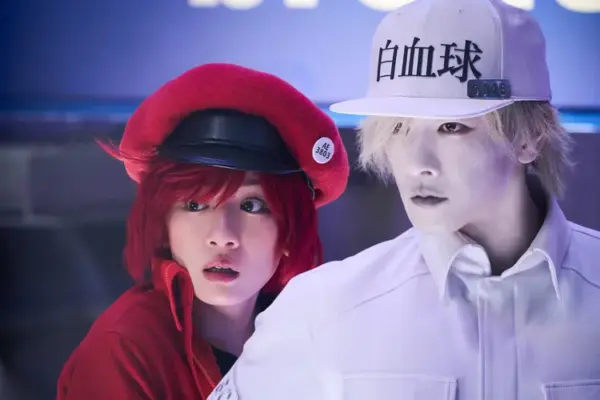 実写『はたらく細胞』の赤血球と白血球キャラクターが並ぶ場面写真。赤いベレー帽と白い帽子の対比が際立つ。