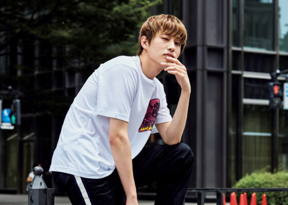 都会の屋外で白いTシャツ姿の若い男性がしゃがみ、顎に手を添えてポーズをとるポートレート（経歴紹介用）