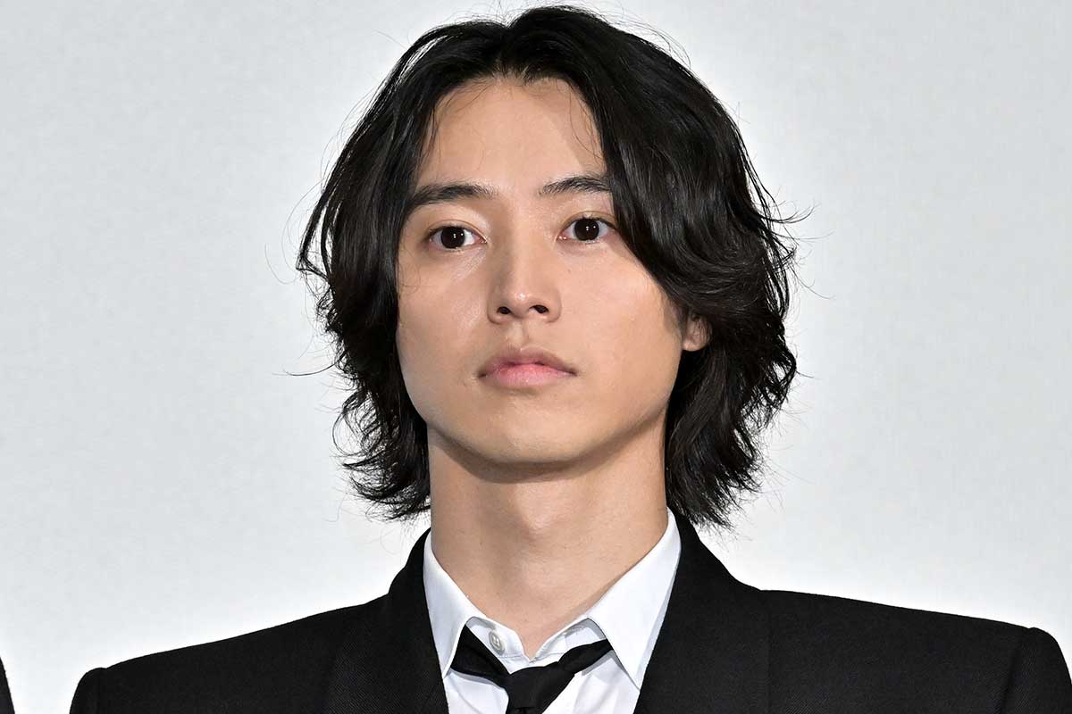 記者会見ステージで正面を向く男性。黒スーツにネクタイの上半身ショット