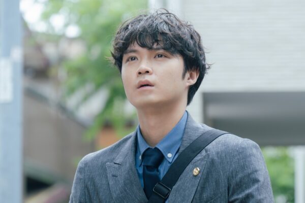 スーツ姿で通勤途中のように外を見上げる磯村勇斗の姿。青いシャツとネクタイを着用し、真剣な表情で立ち止まっているシーン。ドラマ『僕達はまだその星の校則を知らない』の場面写真。