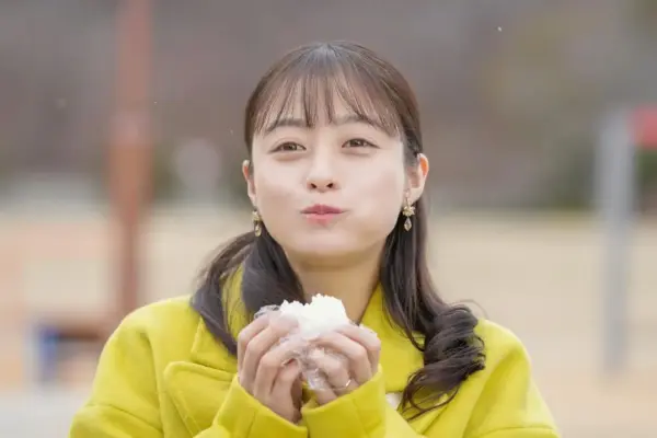 黄色いコートの若い女性がおにぎりを頬張るアップ。朝ドラの温かな日常シーンを切り取ったスチール