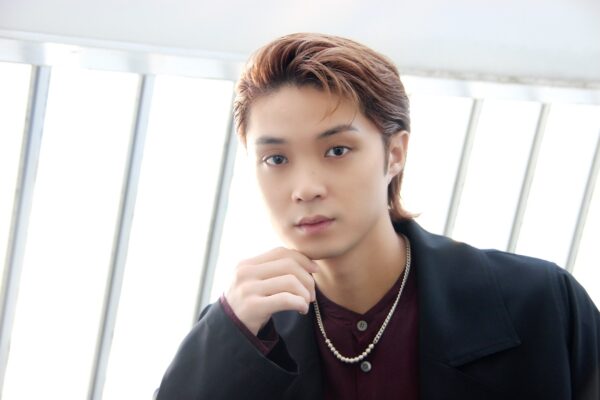 白い背景の前でカメラを見つめる磯村勇斗のポートレート。前髪を上げてネックレスを着け、落ち着いた表情をしている。