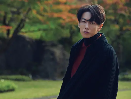 秋の公園でコート姿の男性俳優が静かに佇むポートレート｜大人の雰囲気が漂う近年の活躍と年収アップのイメージ