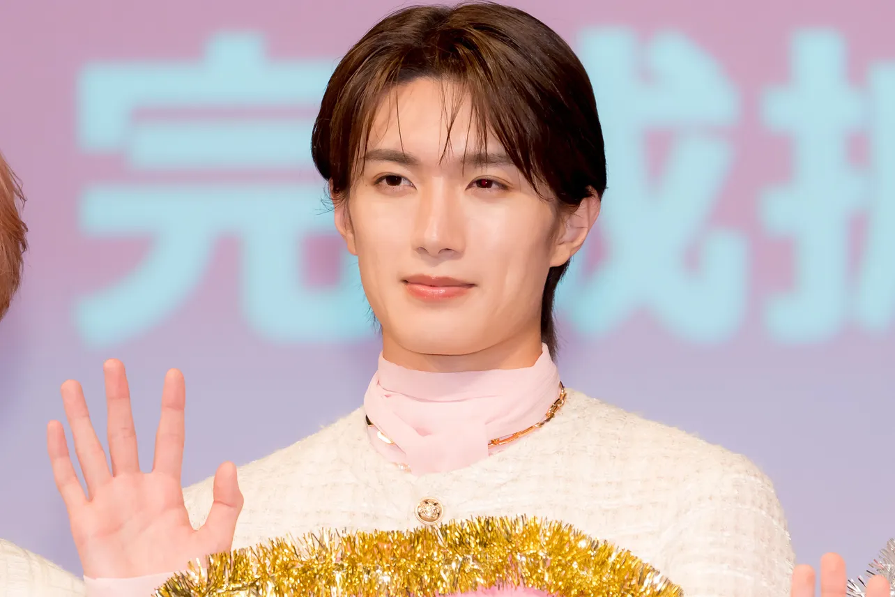 イベント会場で白い衣装の若い男性が手を上げて挨拶する記者会見風の写真（アイキャッチ画像）