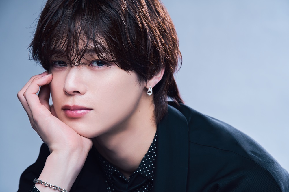 若い男性が頬杖をつきながらカメラを見つめているクローズアップ写真を使った、&TEAM K（古賀祐大）の年収や収入源を解説する記事向けポートレート画像。柔らかい表情と上品なスタイリングが、人気アイドルとしての存在感と高い年収イメージを強調している。