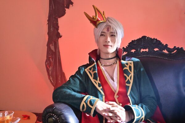 王冠と白髪ウィッグのコスチューム姿の男性が黒い椅子に座るビジュアル写真（特撮・舞台風の衣装）