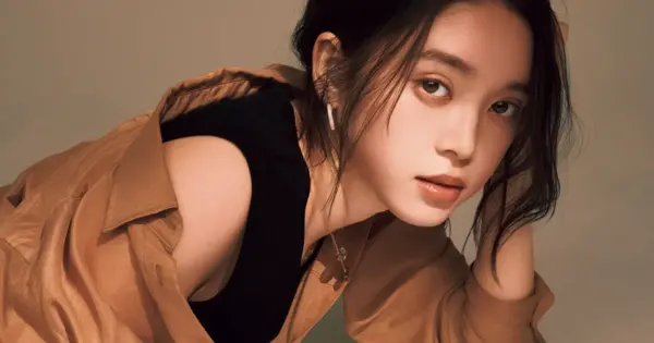 茶色のジャケットを肩に掛けた女性が頬杖をつき、カメラを見る室内ポートレート。柔らかい光で肌の質感が際立つ