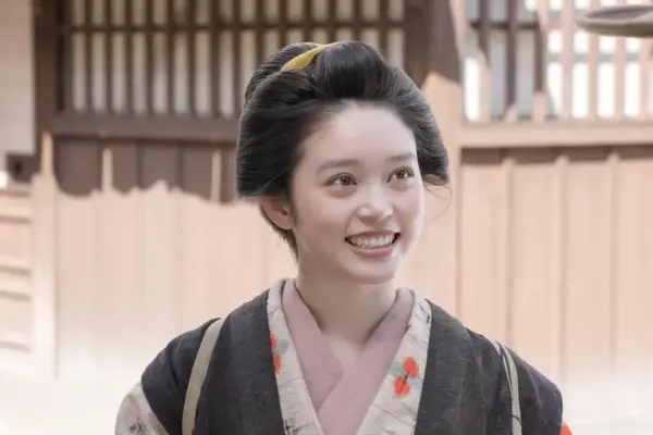 髪をまとめた着物姿の女性が、木造の建物を背景に笑顔で立つ写真。襟元や柄が見える上半身の構図になっている