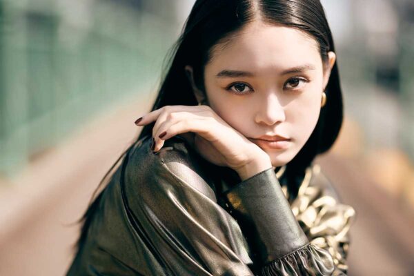 屋外の通路で光沢のあるアウターを着た女性が腕を組み、正面を見つめる写真。背景には柵や建物がぼけて写っている