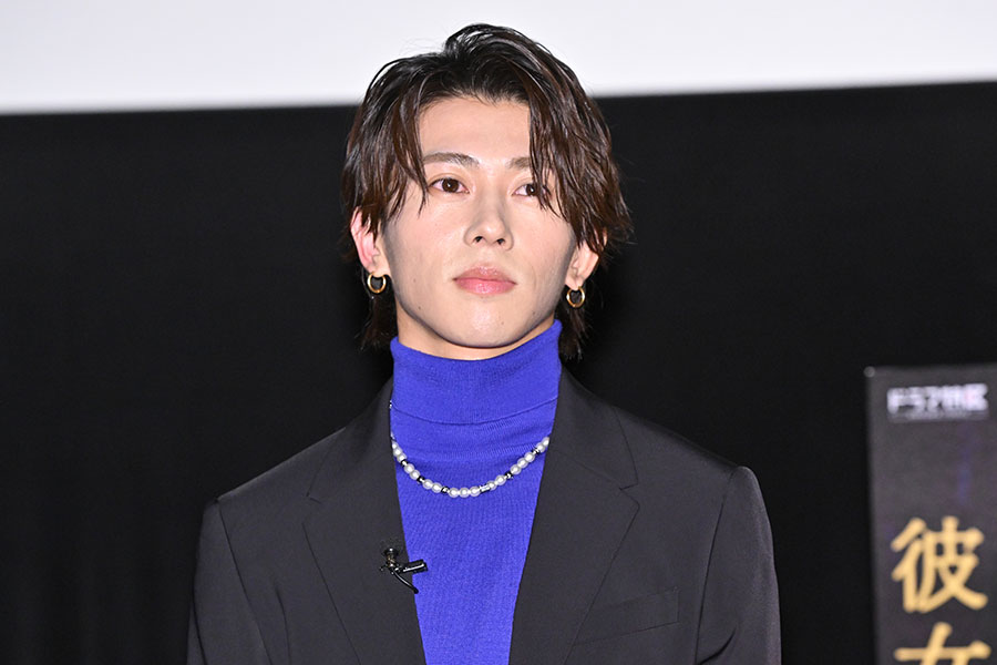 映画の記者会見でスポットライトを浴びる日本人男性俳優が、ブルーのスーツとタートルネックを着て登場。シンプルな背景が彼の外見に焦点を当てています。