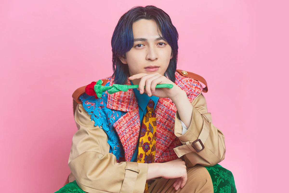 ピンク背景でトレンチコート風の衣装を着た若い男性がバラを持ち、顎に手を添えて見つめるアイキャッチ用ポートレート