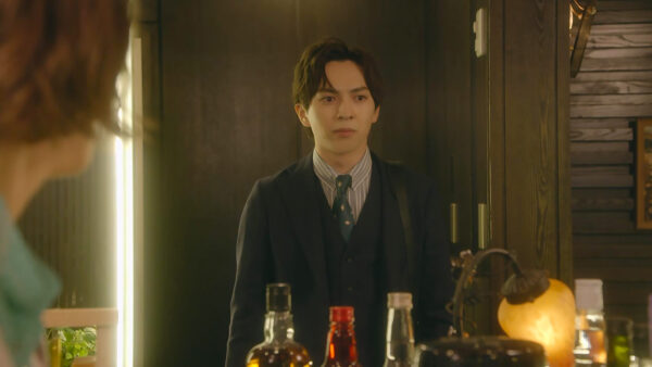 木目調の室内でスーツ姿の若い男性が立ち、手前の人物と向き合うドラマのワンシーン画像