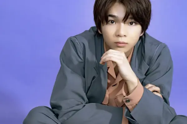 紫の背景でグレーのスーツ姿の若い男性が顎に手を添えて見つめるポートレート写真（経歴記事用）