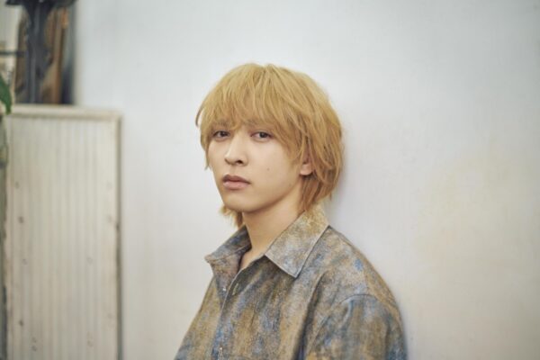 金髪の若い男性が白い壁にもたれて見つめるポートレート写真（経歴紹介記事用）